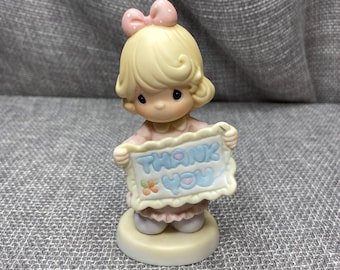 May include: A porcelain figurine of a girl with blonde hair and a pink bow holding a sign that says "Thank You".