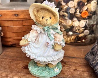Könnte beinhalten: Eine beige Teddybär-Figur mit einer cremefarbenen Haube mit kleinen violetten Blumen, einem gerüschten weißen Kleid mit langen Ärmeln und einem salbeigrünen Band am Hals. Der Bär steht auf einem runden, strukturierten grünen Sockel mit einer weißen Blume.
