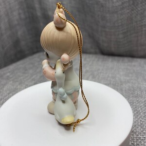 May include: A porcelain Christmas ornament featuring a girl with blonde hair holding a white duck. The ornament is decorated with pink, blue, and white details. The ornament has a gold string for hanging.