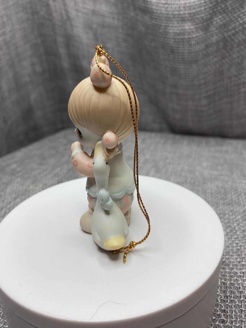 May include: A porcelain Christmas ornament featuring a girl with blonde hair holding a white duck. The ornament is decorated with pink, blue, and white details. The ornament has a gold string for hanging.