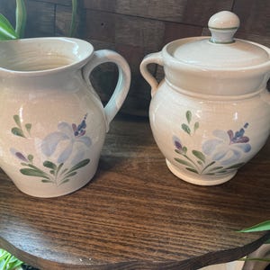 May include: Cream-colored ceramic pitcher and lidded sugar bowl set. Both pieces feature hand-painted floral designs in blue, purple, and green. The pitcher has a handle and spout, while the sugar bowl has two handles and a knob on the lid.