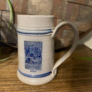 Pode incluir: Uma caneca de cerâmica bege com uma alça curva. A caneca apresenta um design azul e branco com o texto "Twelfth Annual Cambridge Pottery Festival 2003". A caneca tem formato cilíndrico e base larga.