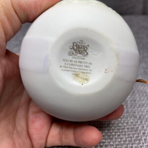 May include: White porcelain Christmas ornament with a gold trim and the words "The Enesco Collection Precious Moments" and "You're as pretty as a Christmas tree" printed on the front. The ornament is made in Thailand.