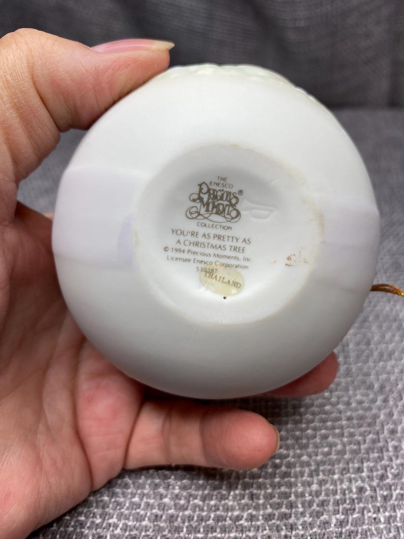 May include: White porcelain Christmas ornament with a gold trim and the words "The Enesco Collection Precious Moments" and "You're as pretty as a Christmas tree" printed on the front. The ornament is made in Thailand.