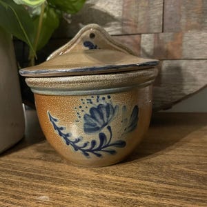 Puede incluir: Un tarro de cerámica marrón y azul de pared con tapa y diseño floral. El tarro tiene un diseño floral azul con contorno blanco.