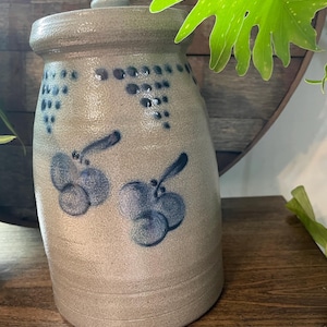 Puede incluir: Una olla de barro gris con tapa, decorada con uvas pintadas de azul y un patrón de puntos azules. La olla está sobre una superficie de madera.