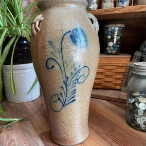 Puede incluir: Un jarrón alto de gres marrón con diseños florales azules y verdes. El jarrón tiene dos asas y una superficie texturizada.