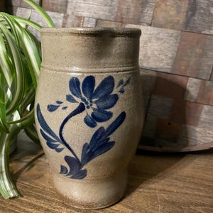 Puede incluir: Una jarra de gres con un diseño floral azul. La jarra está esmaltada en un color marrón claro y tiene una forma ligeramente redondeada.