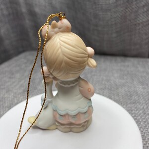 May include: A porcelain figurine ornament of a girl with blonde hair wearing a blue and pink dress holding a white goose. The ornament has a gold string for hanging.
