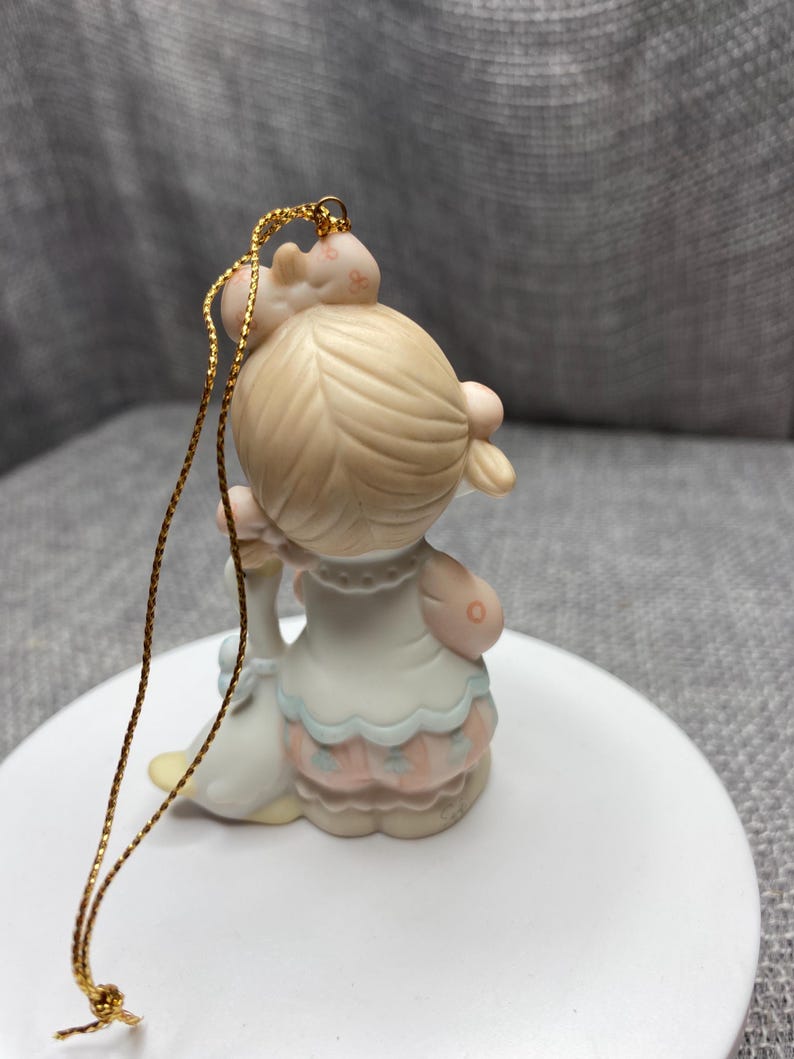 May include: A porcelain figurine ornament of a girl with blonde hair wearing a blue and pink dress holding a white goose. The ornament has a gold string for hanging.