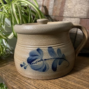 Puede incluir: Un tarro de gres beige con tapa y asa, decorado con diseños florales azules. El tarro se encuentra sobre una estantería de madera, mostrando su encanto rústico. El diseño presenta una superficie texturizada y una estética vintage clásica.