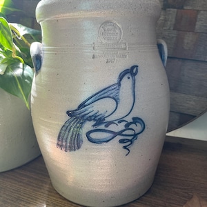 Puede incluir: Una olla de cerámica blanca con un diseño de pájaro pintado en azul y negro. La olla tiene dos asas y un sello que dice "Lowe Pottery Works, Handmade, Johnstown, PA, 1987".