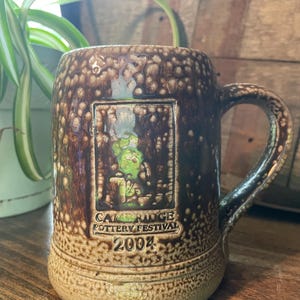 Pode incluir: Uma caneca de cerâmica marrom com um esmalte salpicado. Apresenta uma imagem retangular de uma cena de floresta e o texto "Cambridge Pottery Festival 2004".