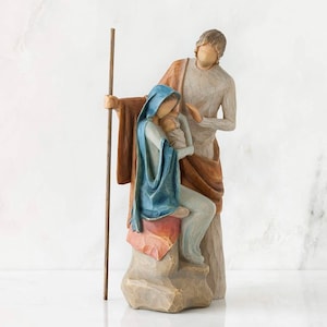 May include: A carved Nativity scene figurine. The sculpture shows Mary in a blue shawl holding baby Jesus, Joseph, and a staff. The figures are in neutral tones with accents of blue, brown, and pink.