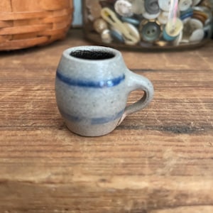 Puede incluir: Una pequeña jarra de cerámica gris con una franja azul alrededor de la parte superior y un asa. La jarra está sobre una superficie de madera.