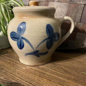 Puede incluir: Un tarro de cerámica beige con tapa y asa, decorado con diseños florales azules. El tarro está sobre una estantería de madera. La tapa tiene un pequeño pomo redondo marrón.