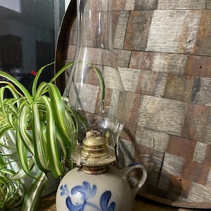 Puede incluir: Una lámpara de aceite vintage con una chimenea de vidrio transparente y una base de cerámica. La base es de color gris claro con diseños florales azules. La lámpara tiene un quemador color latón y un asa pequeña.