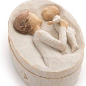 May include: Oval-shaped, off-white decorative box with a lid. The box features a carved design of a person holding a baby. The figures have light brown hair and are dressed in white. The box is a sentimental keepsake.