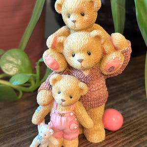 May include: A figurine of three teddy bears stacked together. The largest bear wears a brown sweater, while the middle bear is holding the smallest bear, who is wearing pink overalls. A small white bear is also present.