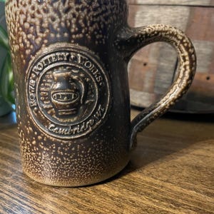 Pode incluir: Uma caneca de cerâmica marrom com uma superfície texturizada e uma alça. A caneca apresenta um emblema em relevo que diz "Rowe Pottery Works, Cambridge, WI."