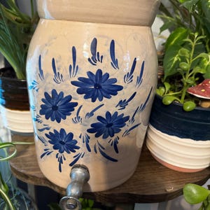 Puede incluir: Un dispensador de bebidas cilíndrico de cerámica beige con motivos florales y de hojas en azul oscuro pintados a mano. El recipiente incluye un grifo metálico fijado en la parte inferior frontal para servir líquidos, con un borde ancho y liso.