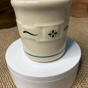 May include: A white ceramic mug with a green floral design. The mug has a green rim and a green floral pattern on the side. The mug is sitting on a white pedestal.