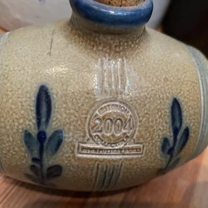 May include: A stoneware bottle with a blue and white floral design. The bottle has a cork stopper and a label that reads "2004".