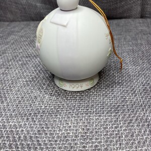 May include: A white porcelain Christmas ornament with a gold string. The ornament has a round shape and features a floral design with the year 1994 printed on the bottom.