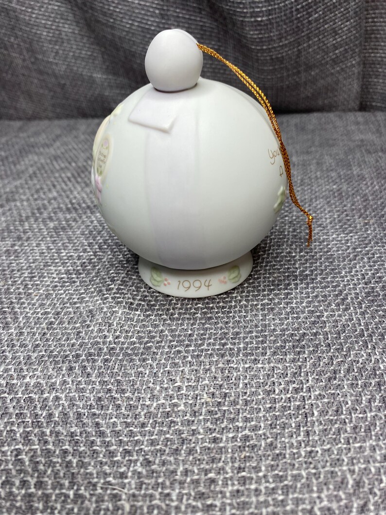 May include: A white porcelain Christmas ornament with a gold string. The ornament has a round shape and features a floral design with the year 1994 printed on the bottom.