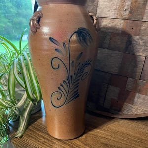 Puede incluir: Un jarrón de gres marrón con un diseño floral azul y verde. El jarrón tiene dos pequeñas asas a los lados.