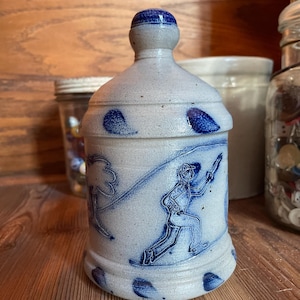 May include: A blue and white ceramic jar with a blue lid. The jar has a design of a person skiing down a hill with a tree in the background.