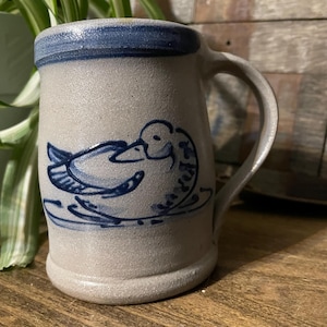 Pode incluir: Caneca de grés com um desenho de pato. A caneca é cinza claro com uma borda e alça azuis. Um pato azul é pintado na lateral. A caneca é de tamanho padrão para café ou chá.