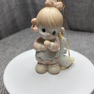 May include: A porcelain figurine ornament of a girl with blonde hair and a pink bow holding a white goose. The ornament has a gold string for hanging.