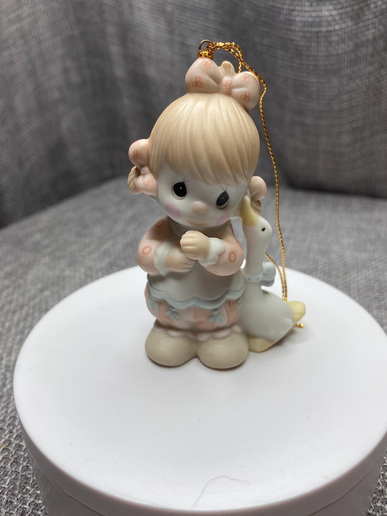 May include: A porcelain figurine ornament of a girl with blonde hair and a pink bow holding a white goose. The ornament has a gold string for hanging.