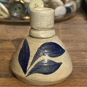 May include: A small, white ceramic vase with a blue leaf design. The vase has a round base and a narrow neck.