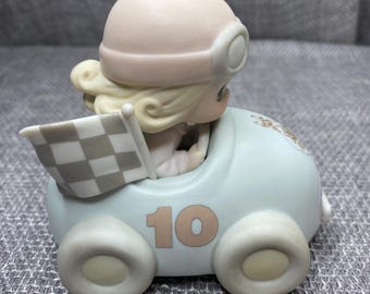May include: A porcelain figurine of a girl wearing a helmet and driving a blue race car with a checkered flag. The car has the number 10 on the side.