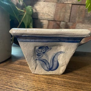 May include: A small, square, stoneware bowl with a blue floral design. The bowl is light gray with a dark blue stripe around the rim and a blue flower illustration on the front. The bowl sits on a wooden surface.