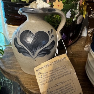 May include: A tan stoneware kerosene bottle lamp with a blue painted heart design and a handle. A white wick protrudes from the top. A beige paper tag is attached, printed with KEROSENE BOTTLE LAMP, usage instructions, and ROWE POTTERY WORKS.
