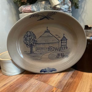 May include: A ceramic plate with a blue and white design of a barn and a tree. The plate is round and has a slightly raised rim.