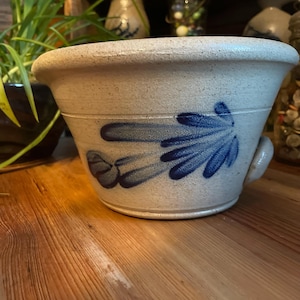 May include: A blue and white stoneware bowl with a blue floral design. The bowl has a handle on one side and is sitting on a wooden surface.