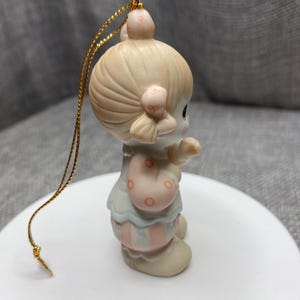 May include: A porcelain ornament of a girl with blonde hair wearing a pink and blue clown costume. She is holding a red and white striped ball. The ornament has a gold string for hanging.