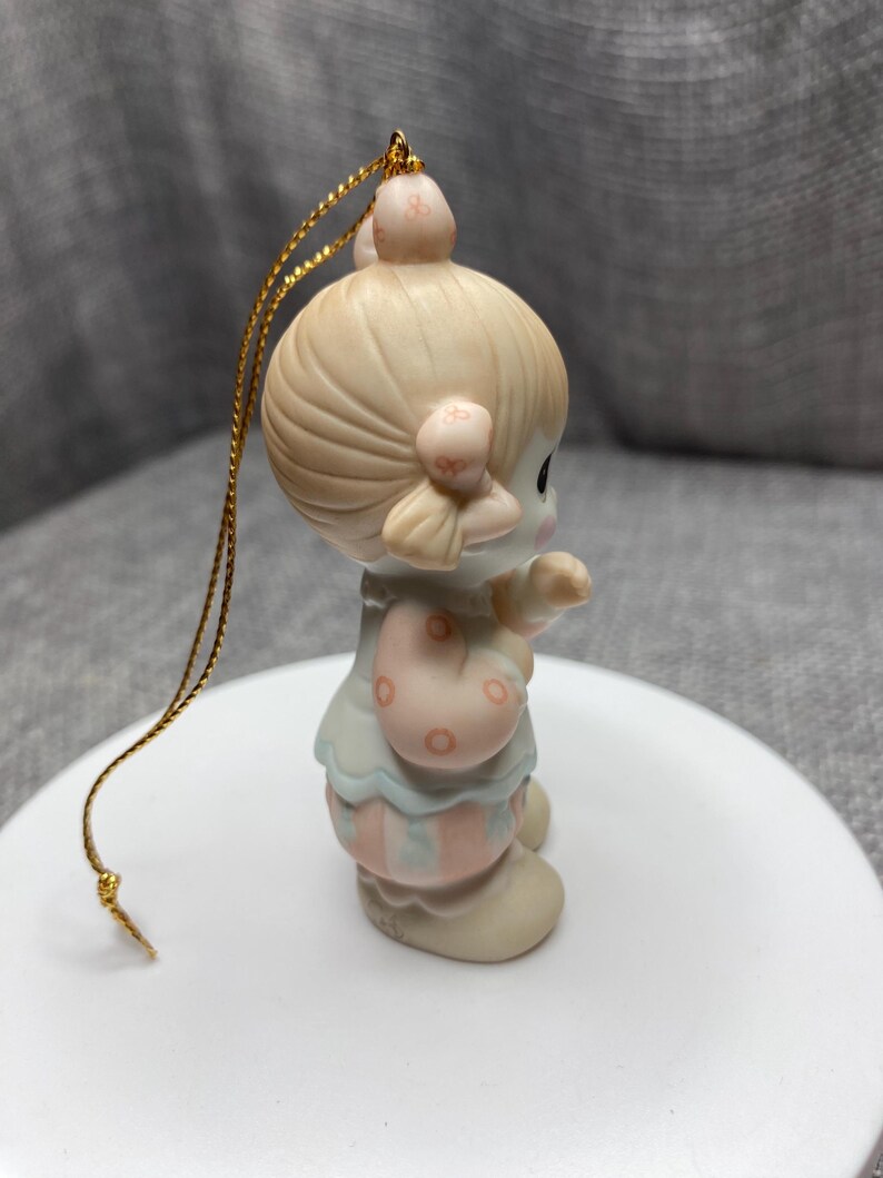 May include: A porcelain ornament of a girl with blonde hair wearing a pink and blue clown costume. She is holding a red and white striped ball. The ornament has a gold string for hanging.
