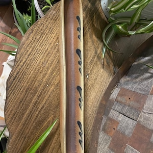 Puede incluir: Una fuente larga de cerámica con forma de artesa redondeada. La fuente tiene un degradado de beige a marrón, con diseños negros en forma de hojas a lo largo del centro. La fuente está colocada sobre una superficie de madera.