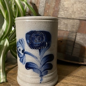 May include: A grey stoneware beer stein with a blue floral design and a blue circular emblem that reads "Hand Made 20 Years 1977-1997".