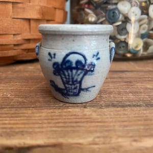 Puede incluir: Un pequeño tarro de cerámica azul y blanco con un diseño de pájaro azul y cesta. El tarro tiene un cuerpo redondeado y una pequeña abertura en la parte superior.