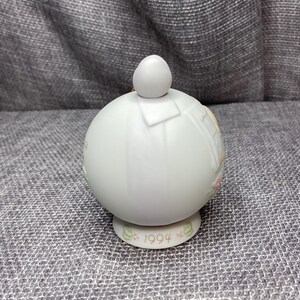May include: White porcelain Christmas ornament with a round shape and a small white ball on top. The ornament features a painted design of a baby angel with wings and a halo. The bottom of the ornament has the year 1994 printed on it.