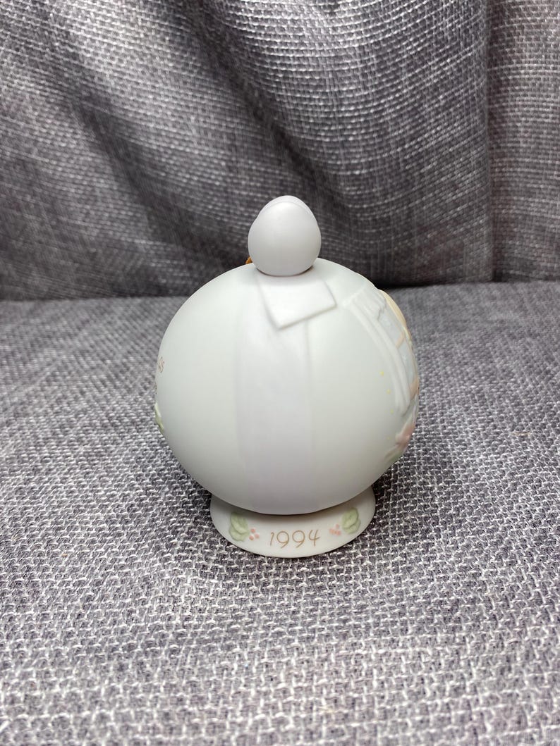 May include: White porcelain Christmas ornament with a round shape and a small white ball on top. The ornament features a painted design of a baby angel with wings and a halo. The bottom of the ornament has the year 1994 printed on it.