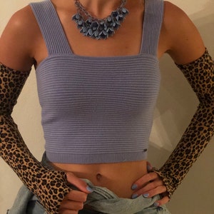 以下が含まれることがあります： ライラック色のリブ編みクロップドトップ、スクエアネック。青い花のペンダントが付いたステートメントネックレスと、ヒョウ柄のアームウォーマーが含まれています。デニムジャケットがウエストに巻かれています。
