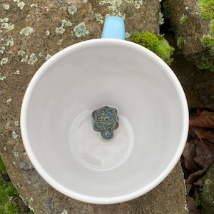 Könnte beinhalten: Weiße Keramiktasse mit hellblauem Henkel und einer kleinen, türkis gefärbten Schildkrötenfigur am Boden. Die Tasse steht auf einer Natursteinoberfläche, mit Moos und Blättern im Hintergrund.