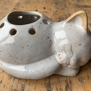 Könnte beinhalten: Eine katzenförmige Vase aus Keramik mit gesprenkelter, cremefarbener Glasur. Die Vase hat mehrere Löcher oben für Blumenstiele. Die Katze liegt mit dem Kopf auf den Pfoten. Ein einzigartiger Dekorationsartikel für zu Hause.